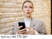 A young woman sporting a trendy hairstyle is playfully taking a selfie against a backdrop of a textured wall, showcasing her modern fashion sense and radiating a cheerful, positive mood. A Young Woman Joyfully and Playfully Posing with Her Phone Against a Textured Wall. Стоковое фото, агентство Ingram Publishing / Фотобанк Лори