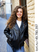A vibrant young woman with curly hair, wearing a chic black leather jacket, poses confidently and effortlessly against a beautifully textured artistic brick wall, greatly enhancing the overall scene. A Smiling Young Woman Wearing a Trendy Leather Jacket Poses Against a Rustic Wall Background. Стоковое фото, агентство Ingram Publishing / Фотобанк Лори