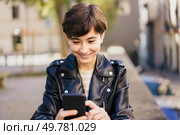 A cheerful young woman wearing a fashionable leather jacket joyfully interacts with her smartphone while outdoors, radiating happiness and a strong connection to her vibrant surroundings. A Cheerful Young Woman Actively Engaged with Her Smartphone While Enjoying the Outdoors. Стоковое фото, агентство Ingram Publishing / Фотобанк Лори