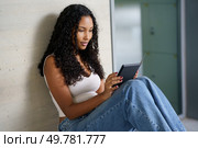 A young woman with beautifully styled curly hair sits comfortably against a chic wall, fully engrossed in her tablet, enjoying a joyful and peaceful moment of leisure and modern technology. A Young Woman Engaged in Using a Tablet in a Relaxed and Cozy Indoor Setting Today. Стоковое фото, агентство Ingram Publishing / Фотобанк Лори