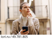 In a charming urban setting, a young woman in an elegant coat and cozy turtleneck focuses on her smartphone, lost in thoughts while enjoying her beautiful surroundings with a sense of peace. A Young Woman Deep in Thought as She Engages with Her Smartphone in a Bustling Urban Setting. Стоковое фото, агентство Ingram Publishing / Фотобанк Лори