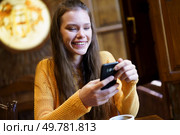 A young woman in a cozy caf smiles while texting on her smartphone. Warm lighting enhances the inviting atmosphere, perfect for social interactions. Cheerful Young Woman Relaxing and Enjoying a Tasty Coffee while Texting on Her Smartphone. Стоковое фото, агентство Ingram Publishing / Фотобанк Лори