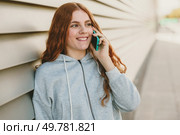A cheerful young woman with long red hair is animatedly speaking on her phone, radiating positive energy while leaning against a stylish wall, ideal for lifestyle and communication themes. A young woman in a grey hoodie smiles while talking on the phone, seated against a wall. Стоковое фото, агентство Ingram Publishing / Фотобанк Лори
