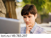 A young woman sits outdoors, surrounded by the beauty of nature, deeply focused on her laptop, embodying concentration and contemplation, as she furthers her studies or works on an important project. A Thoughtful Young Woman Engaged in Serious Work on Her Laptop in an Outdoor Setting. Стоковое фото, агентство Ingram Publishing / Фотобанк Лори