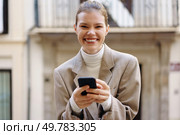 A cheerful young woman wearing a stylish beige blazer smiles broadly while she is engaged in texting on her smartphone, perfectly capturing a delightful moment of joy in a vibrant urban setting. A joyful young woman is smiling brightly while she is texting on her smartphone outdoors. Стоковое фото, агентство Ingram Publishing / Фотобанк Лори
