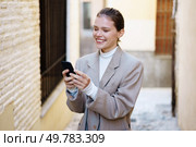 A cheerful young woman dressed in a chic outfit is engaging with her smartphone while walking through a charming alley. A cheerful young woman in a stylish outfit smiles while using her smartphone in a city alley. Стоковое фото, агентство Ingram Publishing / Фотобанк Лори