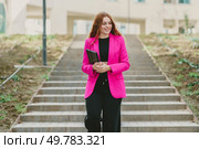 A confident businesswoman in a chic pink blazer ascends outdoor steps with a stylish notebook, embodying professionalism and grace, showcasing her determination to succeed in her career. A Confident Businesswoman Wearing a Vibrant Pink Blazer Poses on the Steps Outside. Стоковое фото, агентство Ingram Publishing / Фотобанк Лори