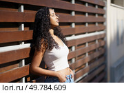 A fashionable young woman with beautiful, curly hair stands gracefully in the warm sunlight, leaning against a rustic wooden wall, radiating confidence, poise, and an aura of serenity and contentment. A portrait capturing the essence of a young woman with curly hair against a wooden background. Стоковое фото, агентство Ingram Publishing / Фотобанк Лори