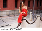 A striking woman dressed in a vibrant and eyecatching red outfit sits elegantly on a pillar chain in a bustling urban setting, exuding an undeniable aura of confidence and unique style. An Elegant Woman Wearing a Stunning Red Fashion Outfit Posing Gracefully Outdoors in Nature. Стоковое фото, агентство Ingram Publishing / Фотобанк Лори