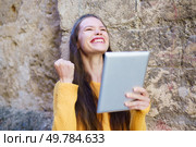 A joyful young woman celebrates her remarkable success while proudly holding up a tablet, expressing her excitement and happiness against the eyecatching backdrop of a rustic wall. A Joyful Young Woman is Celebrating with a Tablet in Her Hand, Radiating Happiness and Enthusiasm. Стоковое фото, агентство Ingram Publishing / Фотобанк Лори