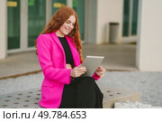 In a charming urban setting, a stylish woman with curly red hair sits outdoors in a pink blazer, engrossed in her tablet, radiating happiness and embodying modern fashion and technology. Stylish and modern woman dressed in a pink blazer, using a tablet outdoors in the city. Стоковое фото, агентство Ingram Publishing / Фотобанк Лори