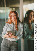 A stylish young woman with long red hair and modern headphones leans against a pillar holding a trendy tablet, embodying creativity and vibrancy in urban life through technology and individuality. A Young Woman Wearing Headphones and Engaging with a Tablet While in a Vibrant Urban Setting. Стоковое фото, агентство Ingram Publishing / Фотобанк Лори