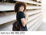 A cheerful young woman with curly hair poses in front of a modern architectural backdrop, exuding confidence and style that captivates everyone, showcasing her vibrant personality. A young woman smiles outdoors with a backpack, embodying a cheerful spirit in a vibrant setting. Стоковое фото, агентство Ingram Publishing / Фотобанк Лори