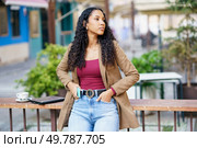 A stylish young woman with curly hair stands outdoors, confidently posing in a chic outfit that highlights her unique fashion style, radiating elegance and selfassurance in a natural setting. A Stylish Young Woman in a Casual Outfit is Posing Outdoors in a Vibrant Urban Environment. Стоковое фото, агентство Ingram Publishing / Фотобанк Лори