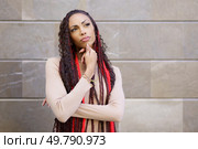 A woman stands thoughtfully against an intriguing textured wall, showcasing her distinctive braided hairstyle adorned with vibrant red accents, embodying confidence, creativity, and personal style. A thoughtful woman sporting braids poses gracefully in a modern urban setting full of life. Стоковое фото, агентство Ingram Publishing / Фотобанк Лори
