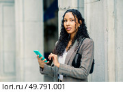 A stylish young woman with curly hair is confidently holding her smartphone against a vibrant city backdrop, showcasing her modern lifestyle and engaging demeanor as she interacts effortlessly. A Stylish Young Woman Engaged in Using Her Smartphone While in an Urban Setting Outdoors. Стоковое фото, агентство Ingram Publishing / Фотобанк Лори