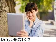 A joyful, bright, and carefree young woman is having a lively video call outdoors, smiling widely while holding a tablet in her hands, surrounded by the beauty of nature all around her. A Young Woman Happily Enjoying a Video Call Outdoors Using Her Tablet in the Sunshine. Стоковое фото, агентство Ingram Publishing / Фотобанк Лори