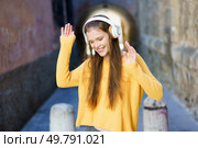 A cheerful young woman dressed in a bright yellow sweater and wearing headphones joyfully dances in a vibrant alley, fully embracing and celebrating the sheer joy and freedom that music brings. A Joyful Young Woman Is Enjoying Listening to Music While Outdoors and Loving Life. Стоковое фото, агентство Ingram Publishing / Фотобанк Лори