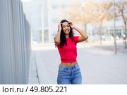 A stylish young woman, sporting beautiful curly hair, poses playfully outdoors in a vibrant red top paired with distressed jeans, perfectly embodying joy, confidence, and a carefree spirit in fashion. A Young Woman is Joyfully Posing Outdoors in a Casual and Trendy Outfit That Radiates Happiness. Стоковое фото, агентство Ingram Publishing / Фотобанк Лори