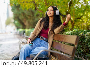 A young woman finds comfort as she relaxes on a bench in a serene park, wearing stylish headphones and deeply immersed in the music, all while being surrounded by the lush greenery of nature. A Young Woman Is Happily Enjoying Listening to Music While Relaxing in a Park on a Sunny Day. Стоковое фото, агентство Ingram Publishing / Фотобанк Лори