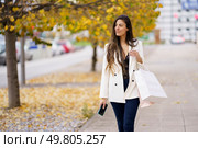 A fashionable woman strolls along a busy sidewalk, holding colorful shopping bags, surrounded by autumn foliage, showcasing her elegance and leisurely lifestyle with a joyful demeanor. A Stylish Woman Delightfully Enjoys Shopping on an Autumn Day Filled with Colorful Leaves. Стоковое фото, агентство Ingram Publishing / Фотобанк Лори