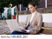 A young businesswoman is sitting on a bench in a bustling urban park, deeply focused on her laptop, embodying the essence of remote work alongside a modern, innovative lifestyle that many aspire to. A young professional is actively engaged in work on a laptop in an urban park setting. Стоковое фото, агентство Ingram Publishing / Фотобанк Лори
