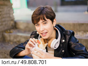 In a lively urban setting filled with energy and movement, a young person sporting a trendy short hairstyle and wearing headphones is happily sitting on the stairs while enjoying a delicious burrito,. A young person joyfully enjoys a delicious burrito while relaxing outdoors in the city. Стоковое фото, агентство Ingram Publishing / Фотобанк Лори