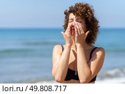Young female in bikini applying sunscreen lotion on face with hands while standing near waving sea during summer weekend. Woman smearing sunblock cream on face near sea. Стоковое фото, агентство Ingram Publishing / Фотобанк Лори
