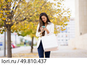 A fashionable young woman checks her phone while strolling with shopping bags on a vibrant autumn day in a lively urban setting, enjoying the surroundings filled with people and nature. Trendy Young Woman Strolling Along a Pathway Carrying Shopping Bags in the Autumn Season. Стоковое фото, агентство Ingram Publishing / Фотобанк Лори