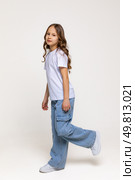 Cheerful genius positive little girl with curly hair in white t-shirt, blue jeans and sneakers has fun and making a step on white background. Happy childhood concept. Copy space. High quality photo. Стоковое фото, фотограф Ирина Михайличенко / Фотобанк Лори