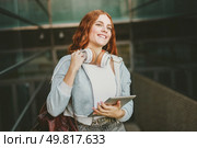 A young woman with long, vibrant red hair, wearing stylish headphones, deeply engrossed in her tablet, enjoying herself in a bustling modern urban environment filled with energy and creativity. A Smiling Young Woman Wearing Headphones and Using a Tablet in a Vibrant Urban Setting. Стоковое фото, агентство Ingram Publishing / Фотобанк Лори