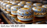 Elegant blue and gold macarons with intricate floral patterns displayed on golden trays in a patisserie, showcasing luxury, craftsmanship and gourmet dessert presentation. Стоковое AI-изображение, AI-художник Катерина Бондарец / Фотобанк Лори