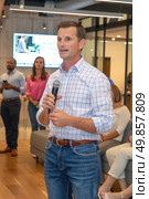 A man stands confidently in front of a diverse crowd, holding a microphone and delivering a speech. Стоковое AI-изображение, AI-художник Данила Большаков / Фотобанк Лори