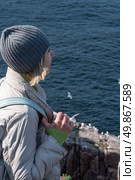 A woman in a gray beanie and light jacket stands on a rocky cliff by the Barents Sea, gazing at the flying seabirds near a nesting colony. Стоковое фото, фотограф Софья Соломонова / Фотобанк Лори