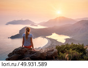 Young woman sitting on the top of rock and looking at the seashore and mountains at colorful sunset in summer. Landscape with girl, sea, mountain ridges and orange sky with sun. Oludeniz, Turkey. Стоковое фото, агентство Ingram Publishing / Фотобанк Лори