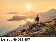 Young sporty man with backpack standing on the top of rock and looking at the seashore and mountains at sunset in summer. Scene with man, sea, mountain ridges and orange sky with sun. Oludeniz, Turkey. Young sporty man with backpack, seashore and mountains. Стоковое фото, агентство Ingram Publishing / Фотобанк Лори