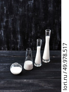Containers of glass cone and spherical shape, filled with white liquid on a dark wooden table on a black background with copy space. Milk saving technology.. Diagonal composition of glass of different shapes and sizes filled with milk or yogurt on a black background. Natural organic dairy product. Стоковое фото, агентство Ingram Publishing / Фотобанк Лори