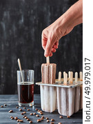 Coffee ice lolly. A woman’s hand takes ice cream with plastic molds, a glass of coffee and coffee beans on a black wooden table. Copy of space. The girl’s hand takes from the mold, homemade natural ice cream on a stick on a black wood table with space for text. Стоковое фото, агентство Ingram Publishing / Фотобанк Лори