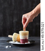 A woman’s hand takes a fruity coffee homemade ice cream lolly in a black plate with ice cubes on a dark wooden background with copy space.. Color berry ice popsicle on a stick in a black plate with pieces of ice on a black wooden background with space for text. Girl’s hand takes ice cream. Стоковое фото, агентство Ingram Publishing / Фотобанк Лори