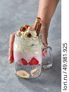 Organic chia seeds with yoghurt, banana, strawberries, cream and wafer tubules in a glass on a gray concrete background. A woman’s hand takes a glass with a healthy dessert. Natural yoghurt with fruits, chia, cream and wafer tubules in a glass. The girl’s hand takes a glass with dessert on a gray concrete background. Стоковое фото, агентство Ingram Publishing / Фотобанк Лори