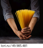 Handful of uncooked spaghetti in the hands of a girl in an apron on black wood vintage table. Mediterranean healthy diet. view bunch of raw spaghetti in female hands on black wood vintage table. Стоковое фото, агентство Ingram Publishing / Фотобанк Лори