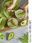 Woman’s hand is holding a freshly healthy green smoothies with kiwas, cucumber and flax seeds, over stone background with asparagus, avocado and wooden board. Vegan meal and detox concept. In female hands a plate with a healthy breakfast and a spoon on the background of a concrete table. Стоковое фото, агентство Ingram Publishing / Фотобанк Лори