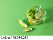 Top view of glass mason jar on a green table with homemade sparkling cocktail with ice, slices of lime, lemon, green mint with plastic straw. Concept of cold alcoholic or non-alcoholic summer drinks.. Refreshing homemade sparkling lemonade with ice in mason jar, slices of lime and lemon, leaf of mint with plastic straws on a green background. Top view. Copy space. Стоковое фото, агентство Ingram Publishing / Фотобанк Лори