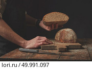 Baker keeps half freshly baked organic bread on a dark background. Hands holding half bread. Стоковое фото, агентство Ingram Publishing / Фотобанк Лори
