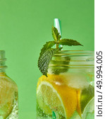 A close-up of a sprig of green mint in focus in a glass jar with cold natural handmade lemonade on green background. Concept of cold alcoholic or non-alcoholic summer drinks.. A sprig of green mint in focus in a glass jar with a refreshing natural handmade cocktail. Citrus fruits slices of lemon and lime, ice, water and plastic straw in the glass. Стоковое фото, агентство Ingram Publishing / Фотобанк Лори