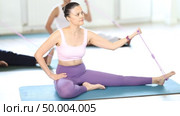 Enthusiastic focused young Asian woman sitting on exercise mat and doing biceps curls with figure eight resistance band to strengthen muscles during group Pilates class in sunlit fitness studio. Стоковое видео, видеограф Яков Филимонов / Фотобанк Лори