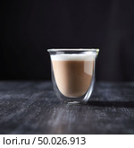 A glass cup of hot flavored cappuccino presented on a black wooden table with copy space.. Freshly brewed cappuccino with foam in a glass cup on a black wooden table around a dark background with copy space. Стоковое фото, агентство Ingram Publishing / Фотобанк Лори