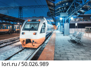 High speed train on the railway station at night in winter. Urban landscape with modern commuter train on the railway platform with illumination at dusk. Intercity vehicle. Passenger railroad travel. Стоковое фото, агентство Ingram Publishing / Фотобанк Лори