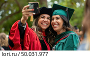 Two jubilant graduates in red and green academic dress capture a selfie to commemorate their graduation day. Стоковое AI-изображение, AI-художник Данила Большаков / Фотобанк Лори