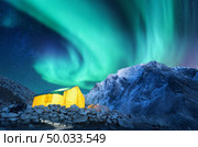 Aurora borealis, yellow glowing tent and snowy mountains. Northern lights, mountain range, starry sky at night in winter. Rocks, sky with stars and polar lights. landscape with green aurora. Space  . Стоковое фото, агентство Ingram Publishing / Фотобанк Лори